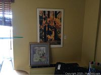 Two framed artworks seen together, one placed on a ledge and the other hanging on the wall.