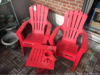 Two red resin Muskoka chairs placed side by side with a small red resin folding table in front of them on a wood tile floor, outdoor setting.