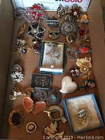 Wide view of assortment of over 25 ladies brooches showing various shapes and colors including floral, bird, animal, and heart designs in metal with rhinestones and enamel.