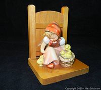 Front and slightly angled view of Hummel book end figurine showing girl in red headscarf with basket of chicks on wooden base against black background.