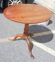 Full view of the round solid walnut table showing the single pedestal base and three curved legs on asphalt surface with vehicle tire in background