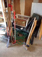 Garden rakes, cable reach tool, spikes, rubber boots, bag, hose, ax, and sledge hammer arranged near bench and on floor.
