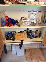 View of cabinet shelf with straps, plastic bags containing hardware fasteners, leather tool belt, handheld electric planer, and two corded drills.