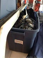 Plastic bin labeled 'Skates' containing black and white hockey skates, hockey helmet with cage, and guards.
