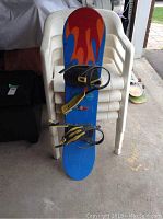 Liquid snowboard standing vertically against white chairs, showing blue deck with red and orange flame graphics and black/yellow bindings attached.