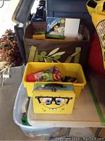 Yellow plastic bin holding various craft items placed in front of a brown fabric tote bag filled with art supplies, and yellow SpongeBob lunchpail at base