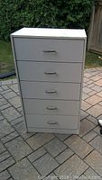 Front view of white 5-drawer dresser with metal handles on a paved outdoor surface.