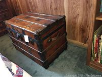 Photo of the chest closed, showing wooden varnished surface, metal reinforcements and latch, on carpeted floor against wood panel wall.