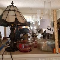 Photo showing Tiffany style lamp with stained glass shade and metal base, alongside three glass kerosene lamps with chimneys and colored bases, ruler for scale.