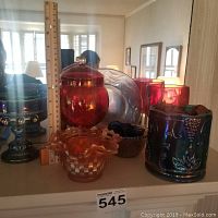 Front view showing various carnival glass pieces including covered candy jars, pedestal compote, ruffled bowls, and decorative containers in deep ruby, blue, and amber colors with iridescent finishes.