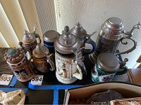 View of 7 assorted German and Austrian beer steins on table showing various designs and pewter lids. Includes castle-shaped and figurative relief steins.