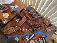 Overview of assorted leather pistol holsters, belts, spurs, and small leather accessories laid out on a table showing condition and engraving details.