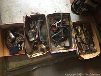 Four cardboard trays containing various hand tools and hardware pieces. View shows assortment of saws, clamps, metal hand tools, and some cables or hoses.