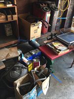 Photo showing red and green metal gas cans with spouts, paint cans, a cardboard box with sprayers, and a red Radio Super Wagon with magazines.