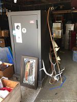 Frontal view of large gray American Security gun vault safe with combination dial and key lock, positioned near boxes and other items in a garage space.