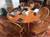 View of entire round wooden table surrounded by seven chairs with various small items on table surface; hardwood floor and kitchen setting visible.