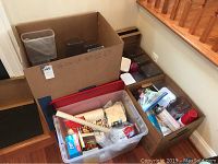 Three cardboard boxes filled with various clear plastic food storage containers stacked inside and on top, along with other disposable food service products in separate boxes and a clear plastic bin with napkins and other paper goods.