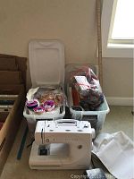 Overview of sewing machine next to two plastic bins containing yarn, ribbons, hooks, and fabric.