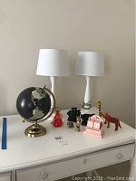 Two white wood table lamps with white shades, one black globe on a brass-tone base, carved wooden animal figures including a tiger, zebra, and a red glass bird, a blown glass cat, and a pink ceramic year calendar on a white dresser.