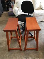 Front view showing the curved seats of the two wooden saddle style barstools and the black office chair behind them.