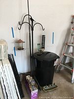 Full view of bird feeder hooks holding two bird feeders, large black trash can, box of birdseed on floor, metal feeder base with four seed cups on floor.