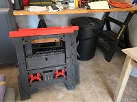 View of black plastic sawhorses with red accents, Black and Decker Workmate workbench behind, black trash can on floor, extension cord on table