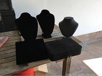 Photo showing 3 bust-style jewelry display stands (2 black velvet, 1 grey), alongside a flat black velvet tray and a large black case with strap on a wooden table.