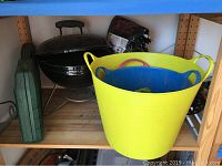 Photo showing portable charcoal grill, two large flexible plastic tubs/garden buckets (yellow and blue), bag of charcoal, against a wooden shelf.