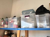 Three clear plastic storage bins on a blue wall-mounted shelf, filled with assorted painting and plumbing supplies