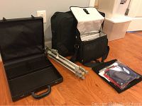 Lot items displayed together: black combination lock briefcase open, aluminum tripod folded, padded black camera bag open, and photo cube in pouch with folded fabric pieces.