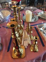 Set of six brass candle sticks and one brass pillar candle holder with white pillar candle and taper candles on a table among other decorative items.
