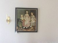 Framed painting of three young girls with one holding a cat, hanging on a wall near a sconce.