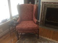 Front view of the pink floral patterned wingback chair with wood arms and legs, placed near a window and fireplace.