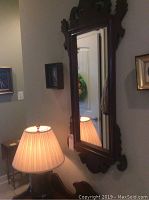 Vertical rectangular mirror with dark carved wood frame and beveled glass reflecting a lamp and coat rack