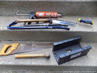 Tools displayed on concrete steps: 2 crowbars, claw hammer, metal hammer, hand saw with yellow handle, plastic mitre box, caulk gun, and utility knife.