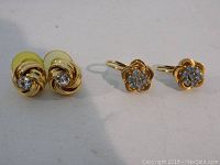 Two pairs of earrings shown: one pair with blue stones in a flower shape, one pair with clear stones in a swirl design.