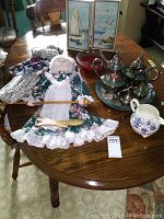 Photo showing silver plated coffee and tea service, wall dolls, framed pictures, red glass bowl, and Royal Staffordshire jug on wooden table.