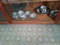 Glass cabinet shelf displaying various Johnson Bros. Friendly Village tea cups, saucers, bowls, and a mug.