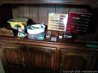 Wide shot of Pyrex casserole dish, two boxed sets of steak knives, ceramic rooster figurine, collector spoons and other kitchen items on wooden cabinet surface
