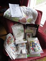 Photo showing embroidery quilt block with colorful hand-stitched birds and floral patterns, cross stitch kit packets, pattern booklets, and embroidery floss packs scattered on a chair.