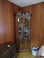 Full view of the corner curio cabinet placed in a wood-paneled corner of a room, showing the curved decorative top, glass upper doors, and wooden lower doors.