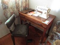 Front view of Singer Stylist model 533 sewing machine mounted in wooden cabinet with stool alongside. Manual and scissors on cabinet top.
