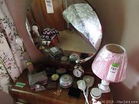 Photo showing vanity set with small mirror, multiple compact trinket boxes, cuff links in a box, small round alarm clock, and small dog figurine on wooden dresser with floral curtain backdrop.