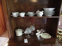 Wide view showing china set stacked on top and glass votive lights and additional items below on shelf