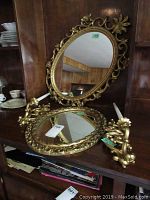 Two gilded oval mirrors with ornate decorative frames placed on a shelf, one is larger, one smaller, reflections visible in mirrors