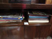 Wide view of two stacks of vinyl record albums on wooden shelf with visible wear on covers.