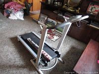 Manual folding treadmill with silver frame and black belt in a carpeted room.