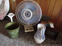 Photo showing an 18 inch electric fan, ceramic pitcher with blue pattern, green wastebasket, and a radio.