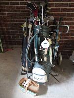Photo showing all three sets of golf clubs in three bags, one green Augusta bag, the others navy blue and black, with some club headcovers visible and a folding push cart behind