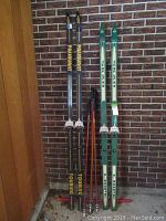 Full view of two pairs of skis: black Passavant Touring skis and green Skan Loipe skis, along with four ski poles leaning against a brick wall.